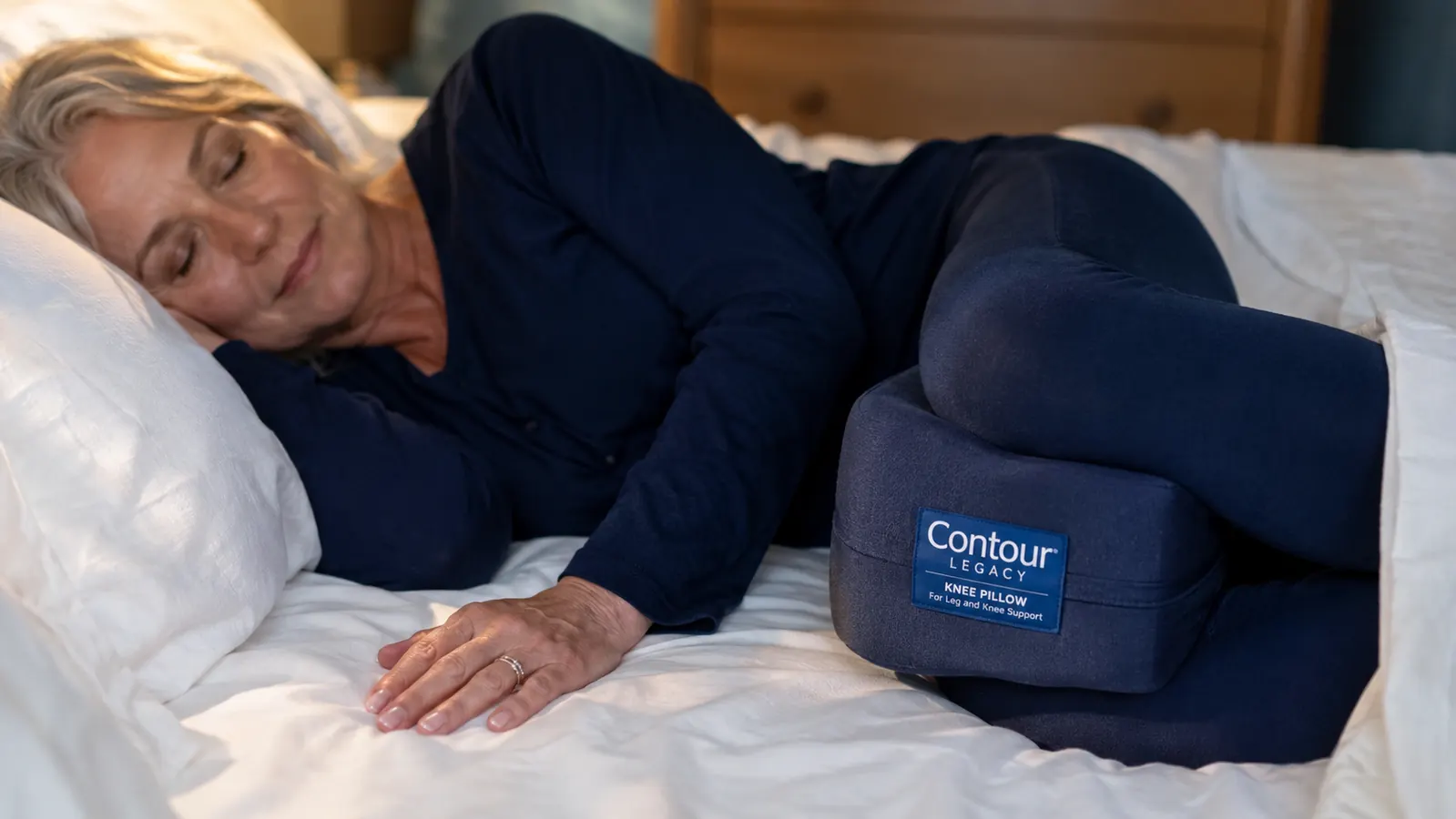 Close-up of a white foam knee support pillow positioned between two bent knees under the covers