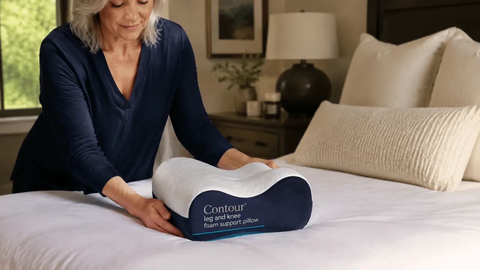 The Contour Legacy knee pillow product placed on top of a neatly made bed with white and cream bedding