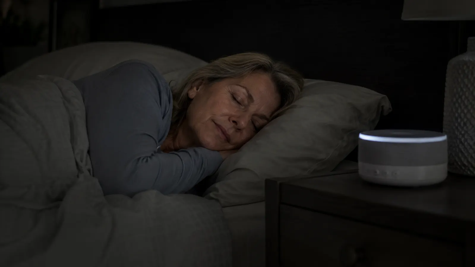 A woman in her late 50s sleeping peacefully in a dark bedroom with a white noise machine glowing softly on the nightstand