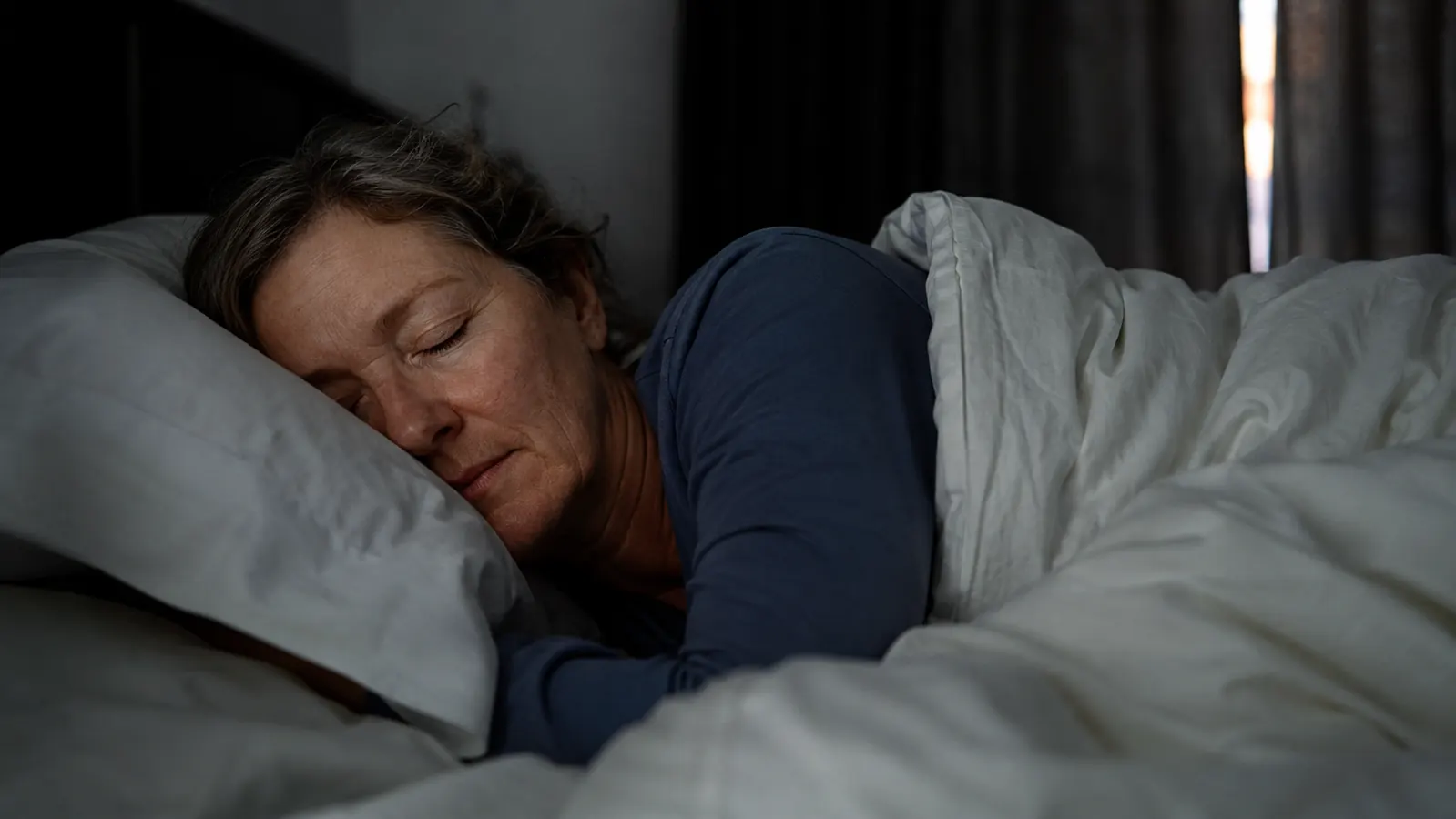 A woman sleeping peacefully in a dark bedroom, white bedding, soft morning light beginning at the curtain edge