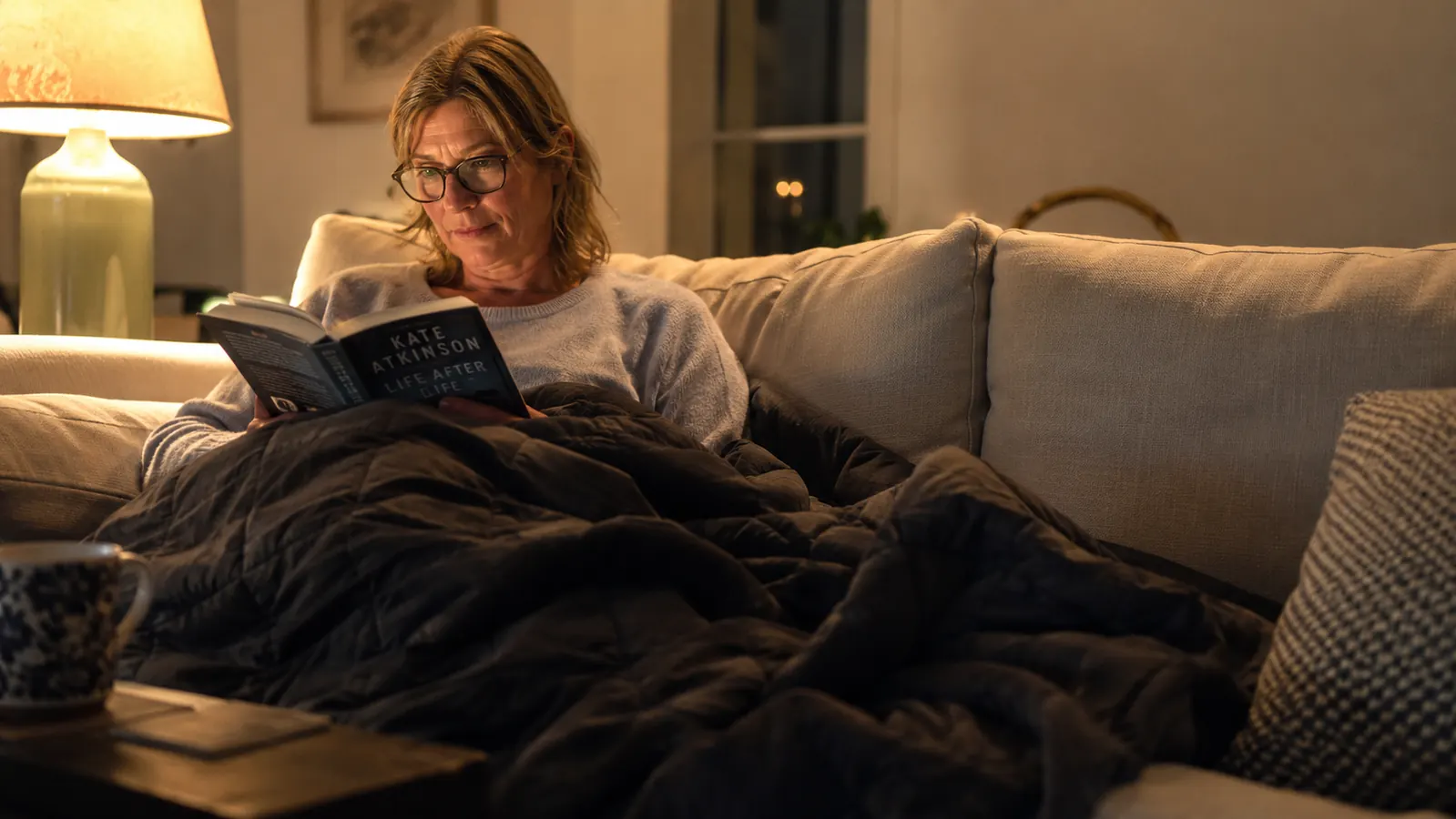Person on a couch reading a book under a weighted blanket, relaxed evening scene with warm lamp light