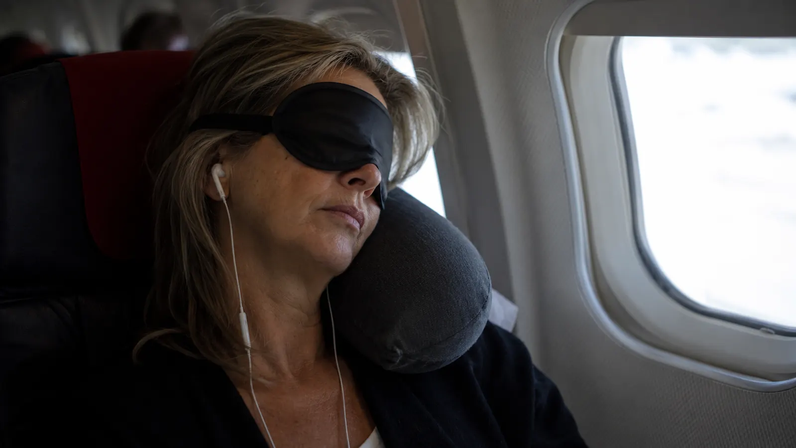 Woman wearing a sleep mask in an airplane window seat, head resting against a travel pillow, eyes closed