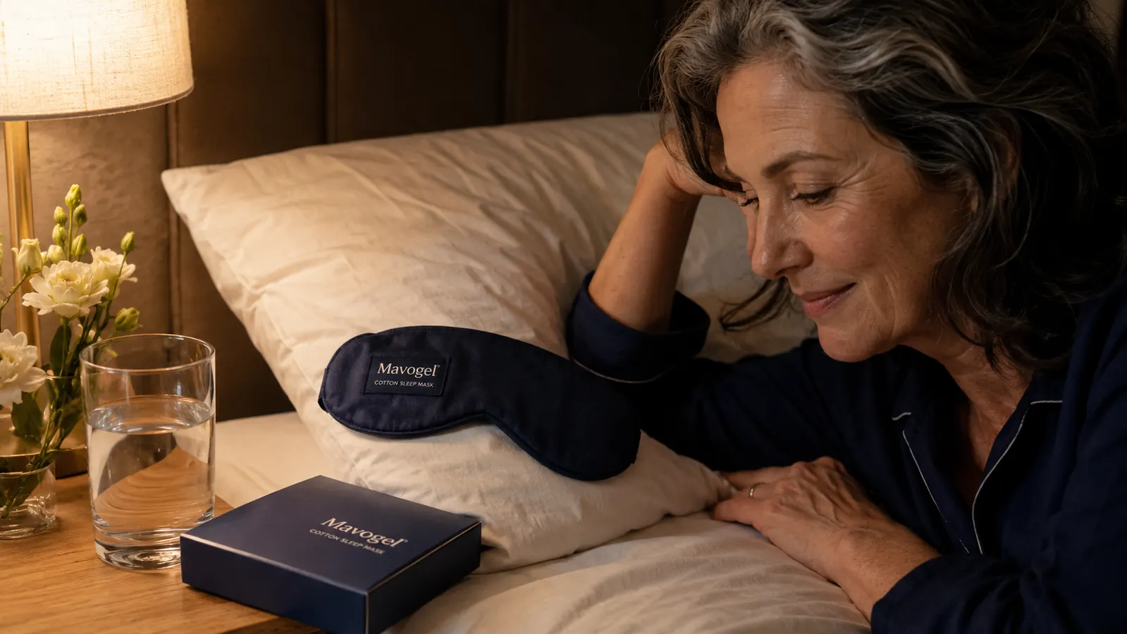 Mavogel sleep mask laid flat on a white cotton pillowcase next to a glass of water on a nightstand